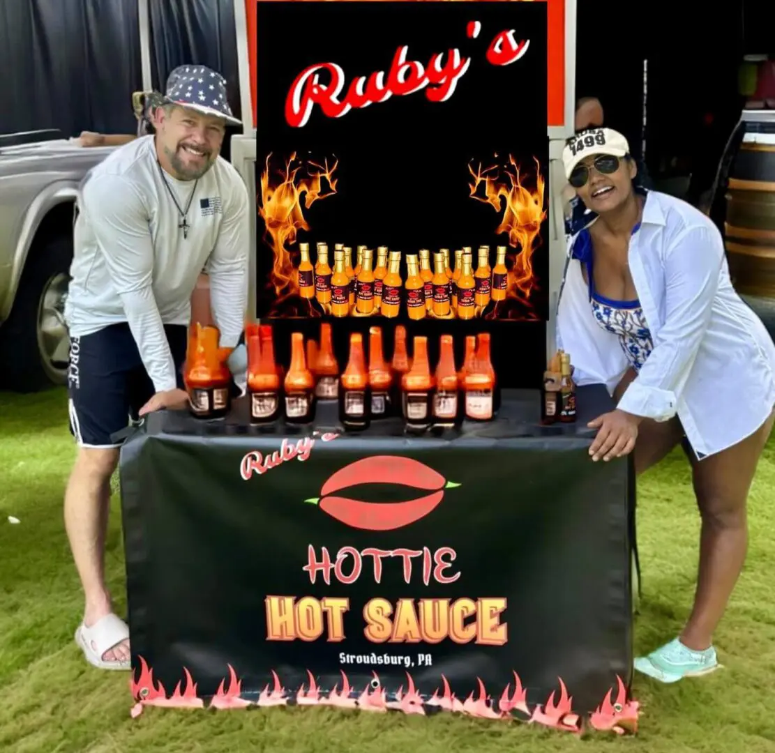 Two people stand behind a display table for Ruby's Hottie Hot Sauce, featuring various hot sauce bottles and promotional signage with flames.