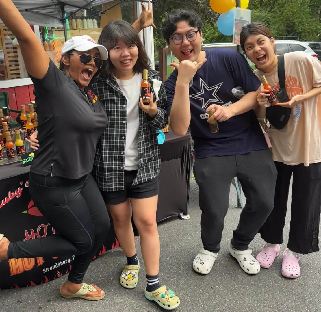 Four people stand together outdoors, smiling and holding bottles. They are in casual attire, and a table with products is visible in the background.