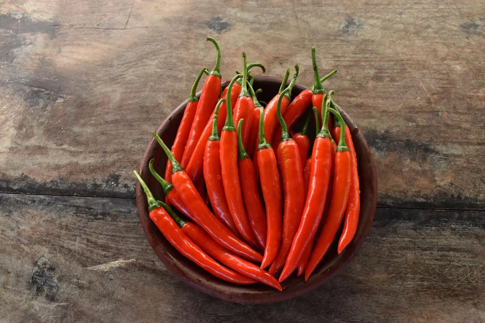 A bowl filled with fresh, red chili peppers sits on a wooden surface.