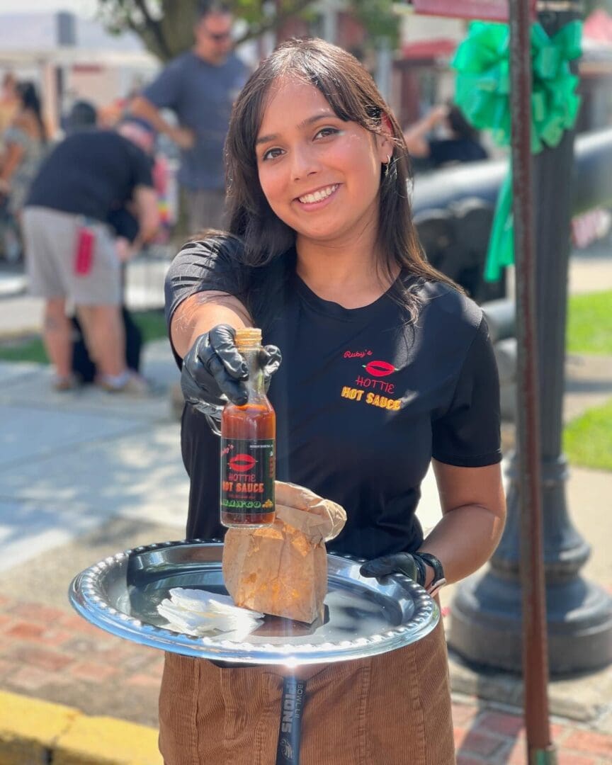 A woman stands outdoors holding a bottle of hot sauce on a tray. She is wearing the Black Ruby's Tee and a glove on one hand, smiling at the camera.